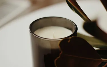 A lit candle in a dark glass holder sits on a white surface. A brown leaf partially obscures the candle, creating a warm, cozy atmosphere.