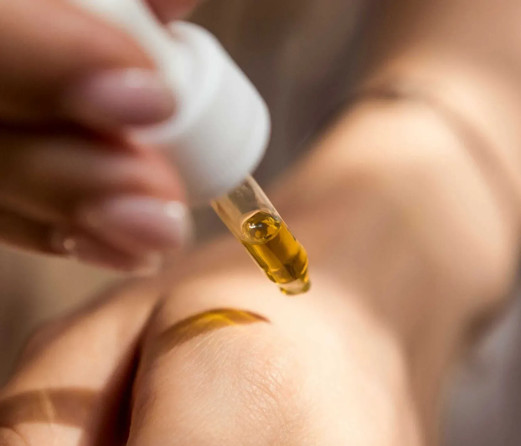 A hand holding a dropper releases yellow oil onto the back of another hand, conveying a sense of care and skincare in warm lighting.