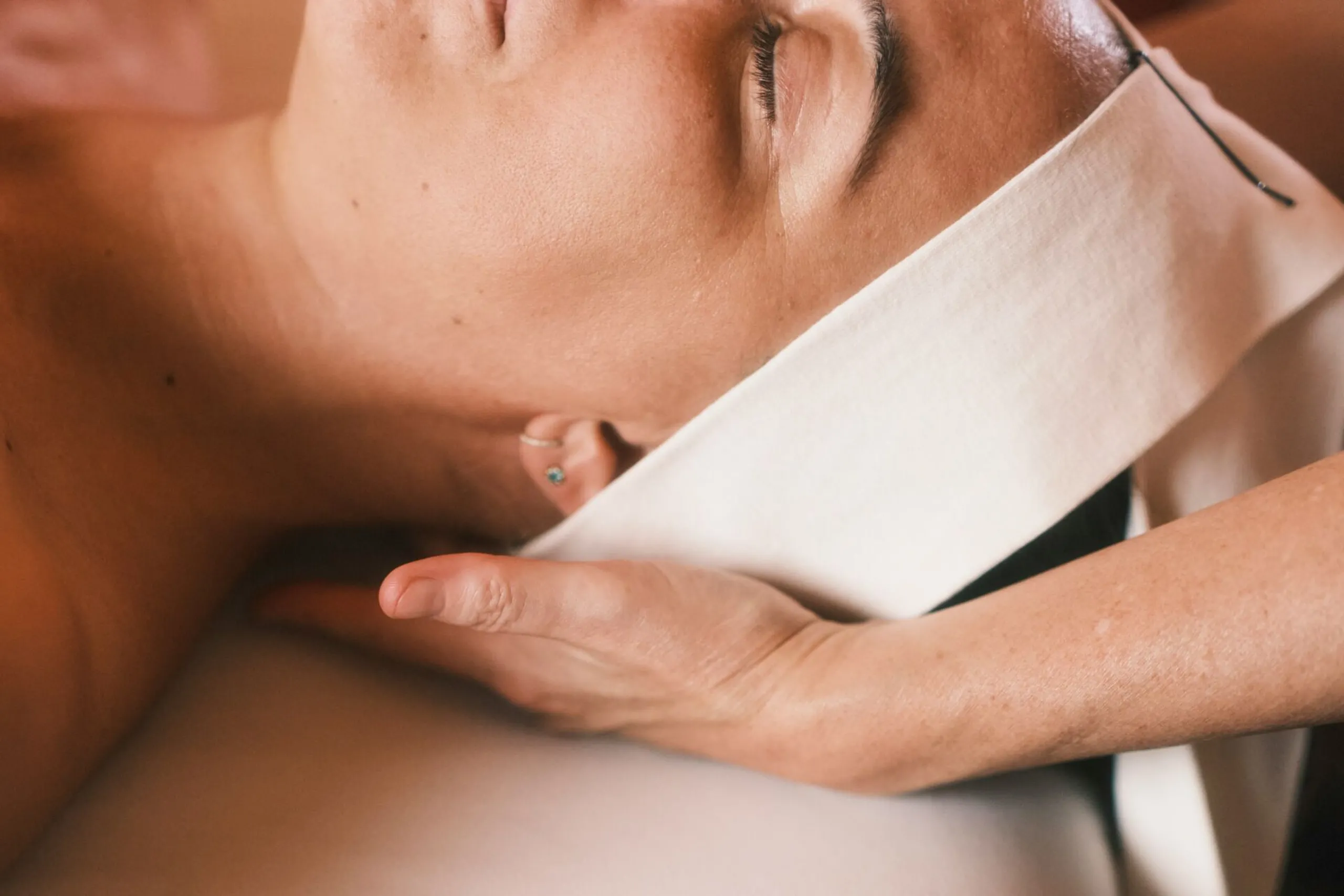A person reclines with eyes closed, receiving a facial massage. Two smooth rose quartz stones are on a cushion, creating a calming, serene atmosphere.