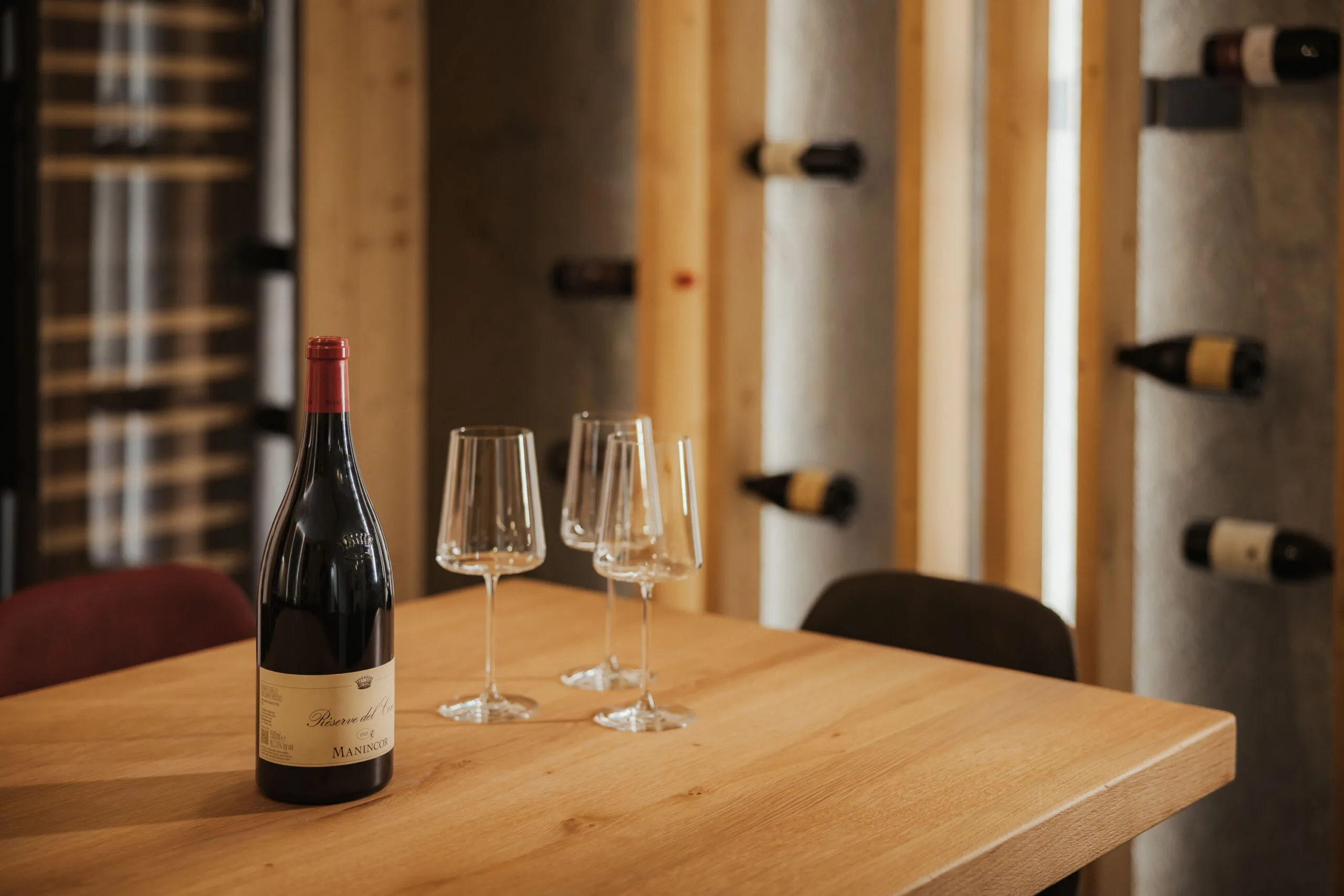 Ein Holztisch mit einer Flasche Rotwein und drei leeren Weingläsern. Im Hintergrund ein Weinregal an einer Holzwand, das eine warme, gemütliche Atmosphäre vermittelt.