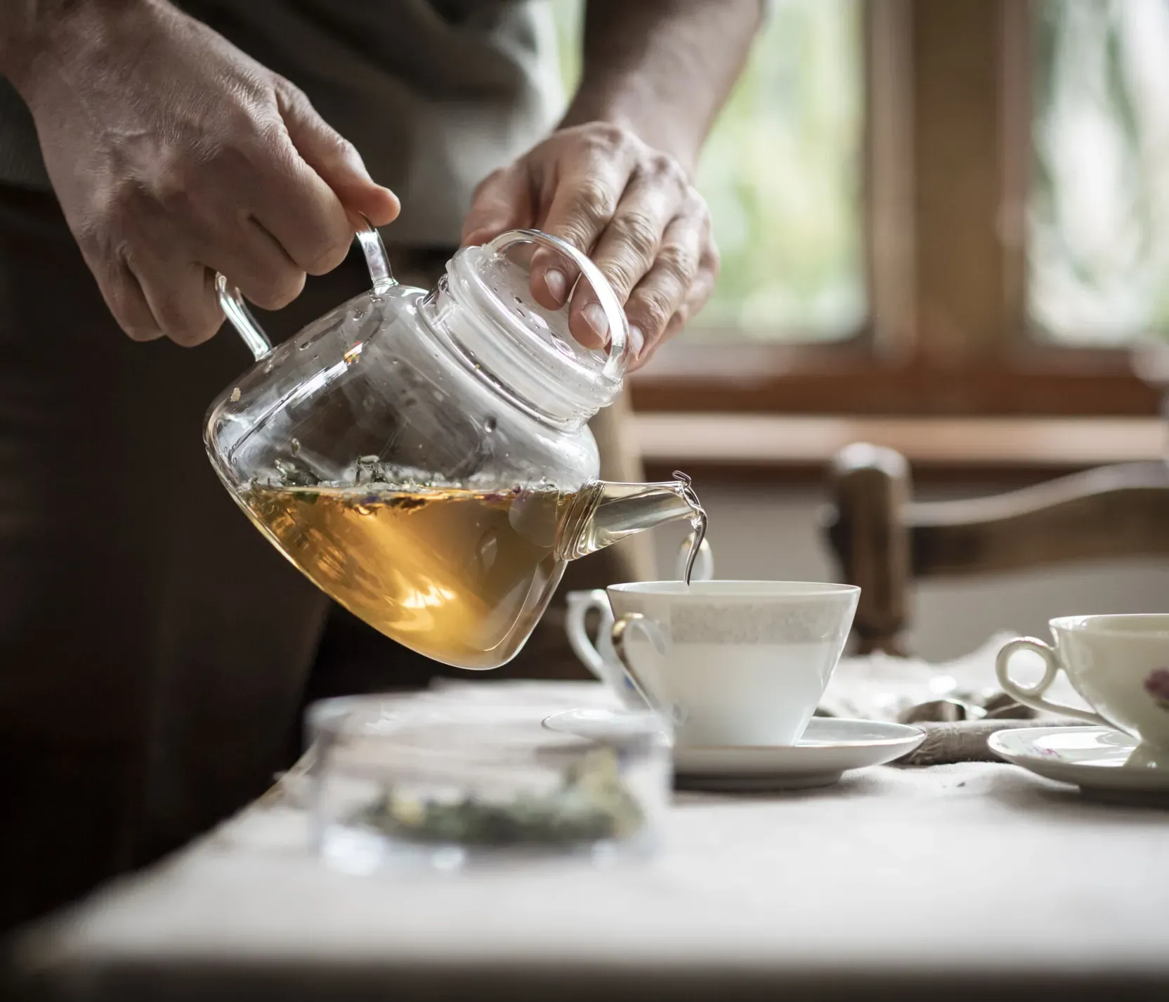Person gießt Tee aus einer Glas-Kanne in eine Tasse auf einem Tisch in einem gemütlichen Raum mit Fenster im Hintergrund.