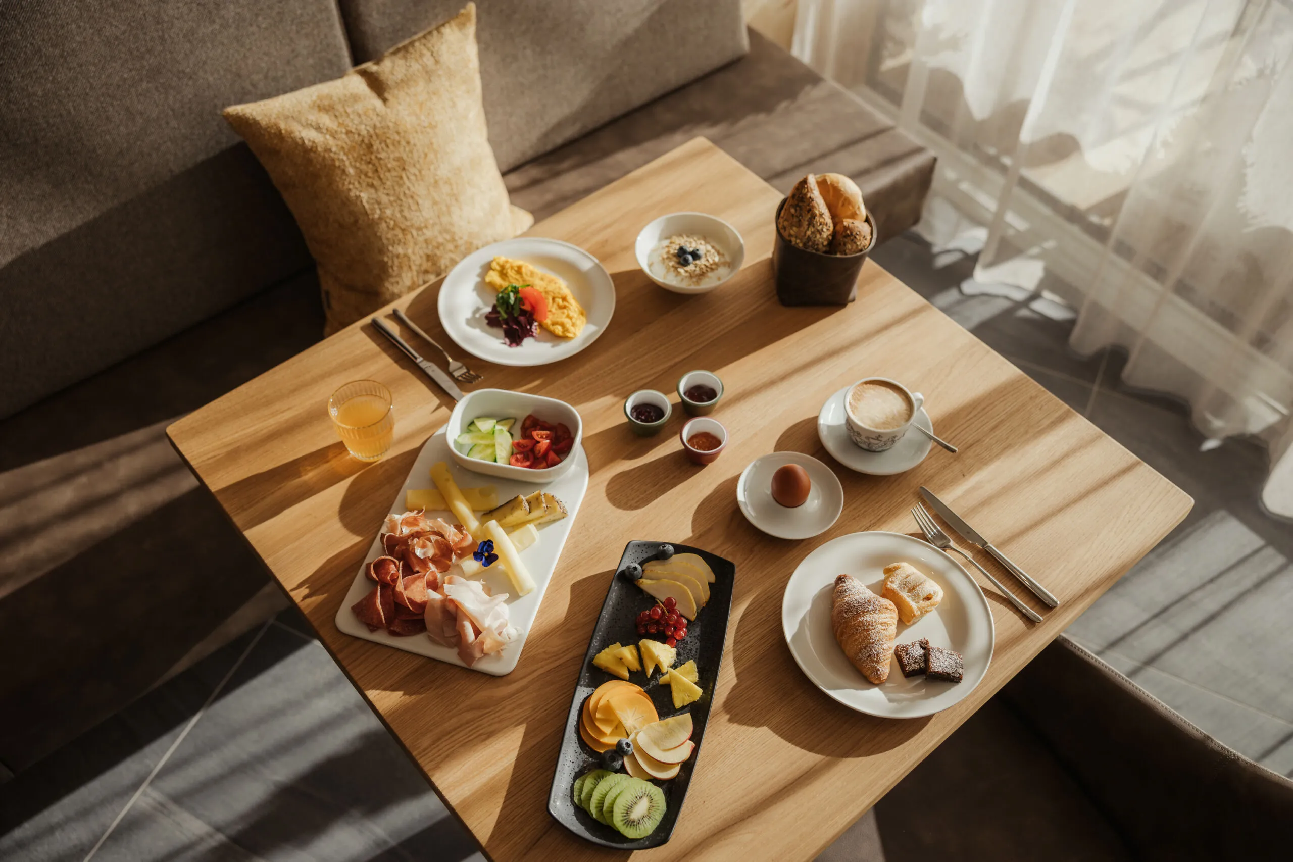 Gemütliche Frühstücksszene auf einem sonnenbeschienenen Holztisch mit Omelett, Obstplatte, Gebäck, Schinken, Käse, Kaffee, Saft und Brot, die Wärme und Vielfalt vermittelt.