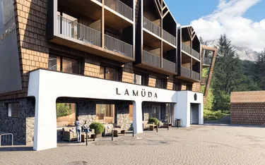 Modernes Hotel La Müda in Alta Badia, Dolomiten, mit Terrasse, Sitzecke, Holz- und Steinfassade, umgeben von Bäumen und Bergen, blauer Himmel.