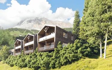 Moderne Gebäude mit Balkonen in grüner Hügellandschaft vor einer Bergkulisse, umgeben von Bäumen, in Alta Badia, Dolomiten.