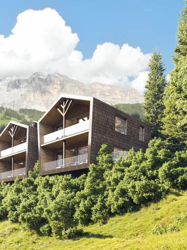 Moderne Gebäude mit Balkonen in grüner Hügellandschaft vor einer Bergkulisse, umgeben von Bäumen, in Alta Badia, Dolomiten.