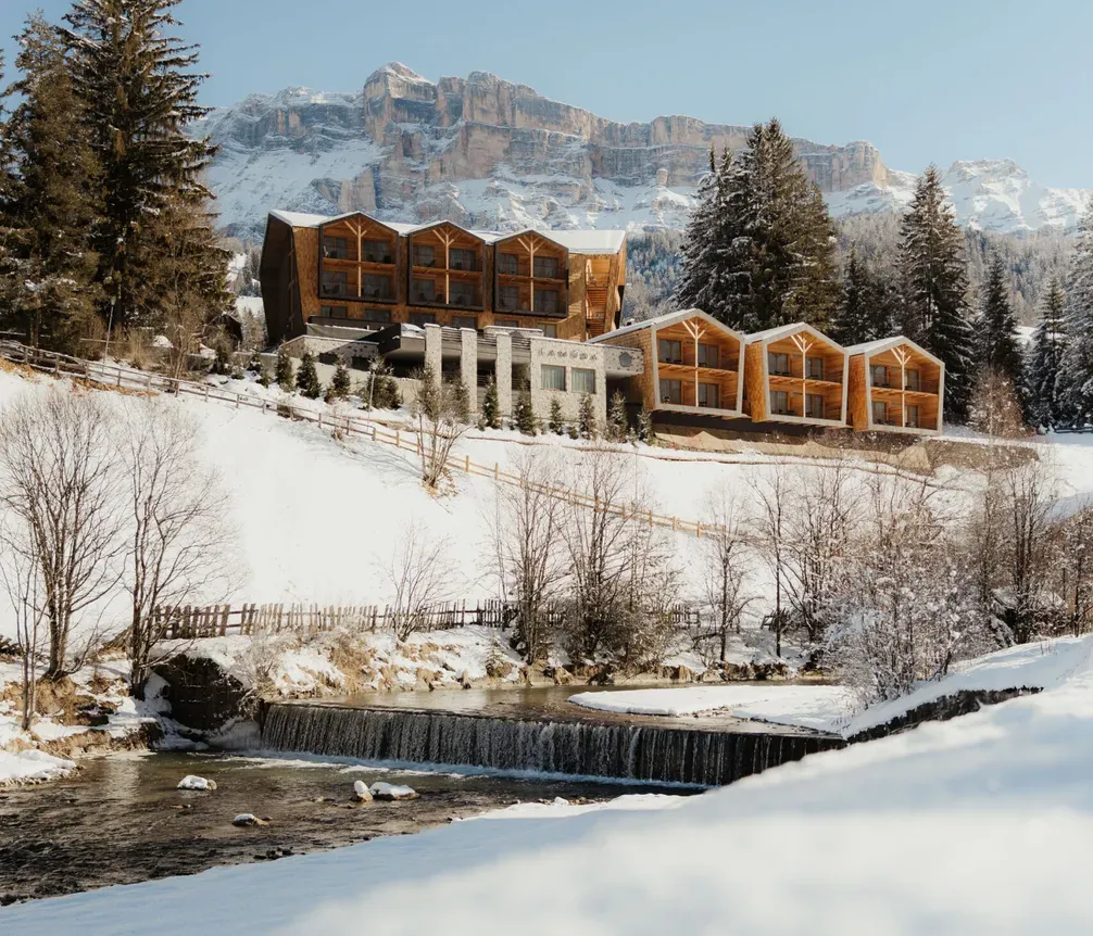 Snowy mountain landscape with wooden lodges surrounded by pine trees. A serene river flows in the foreground, with clear blue skies above. Peaceful winter scene.