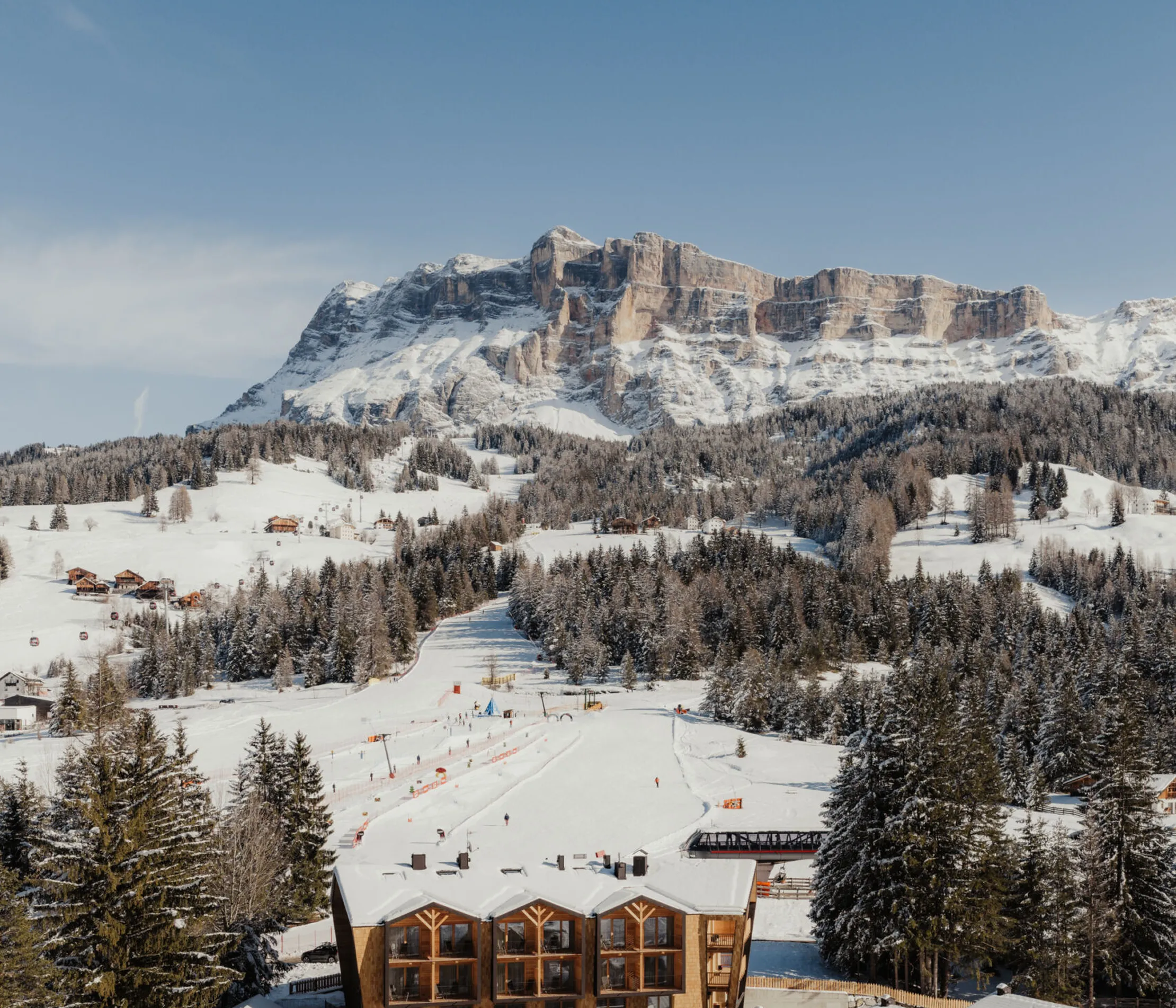 Snowy mountain scene with a ski resort below, rustic wooden architecture surrounded by trees. A majestic snowy peak and blue sky in the background.