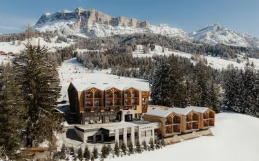 Snowy mountain resort with wooden architecture set against majestic peaks and pine forests. Clear blue sky completes a serene winter scene.