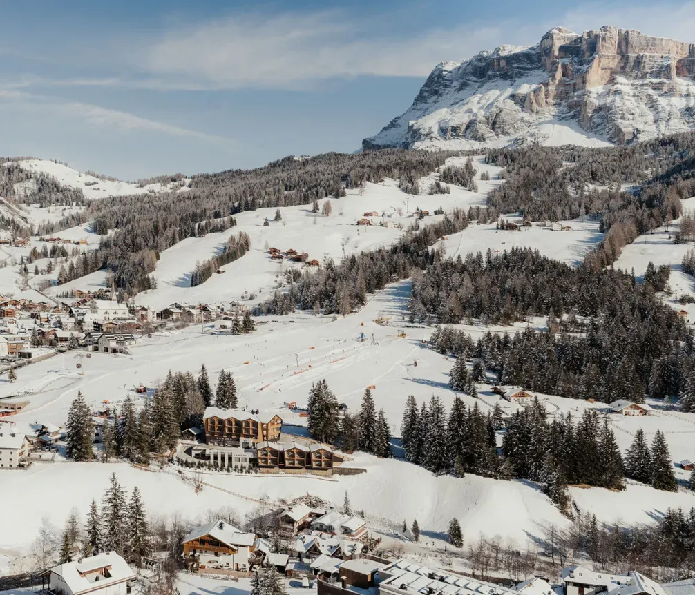 Snowy alpine village nestled among pine forests, with ski slopes winding through the landscape. Majestic mountain peak in the background under a clear blue sky.
