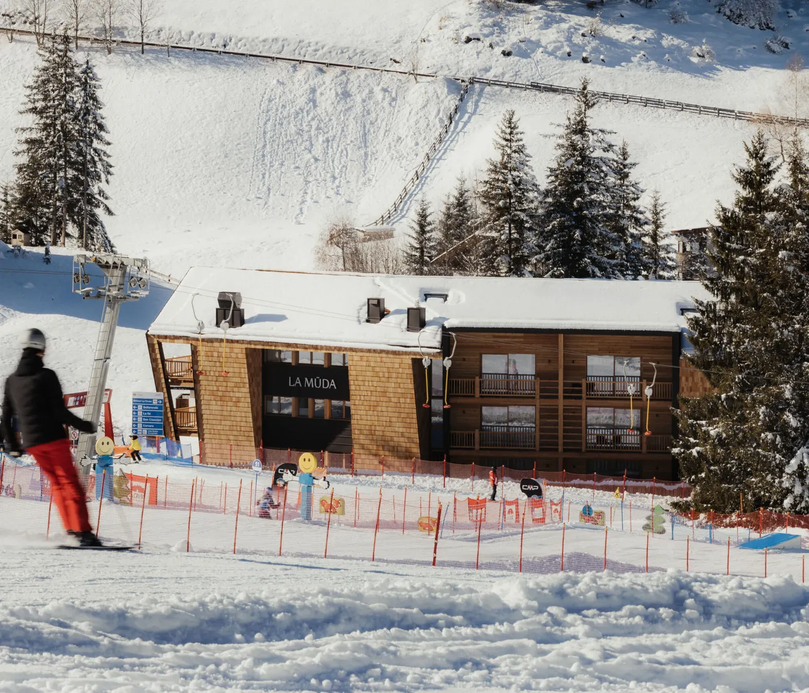 Skier in red pants glides on snow toward a wooden building labeled "La Muda," surrounded by snow-covered trees and a sunny, clear sky.