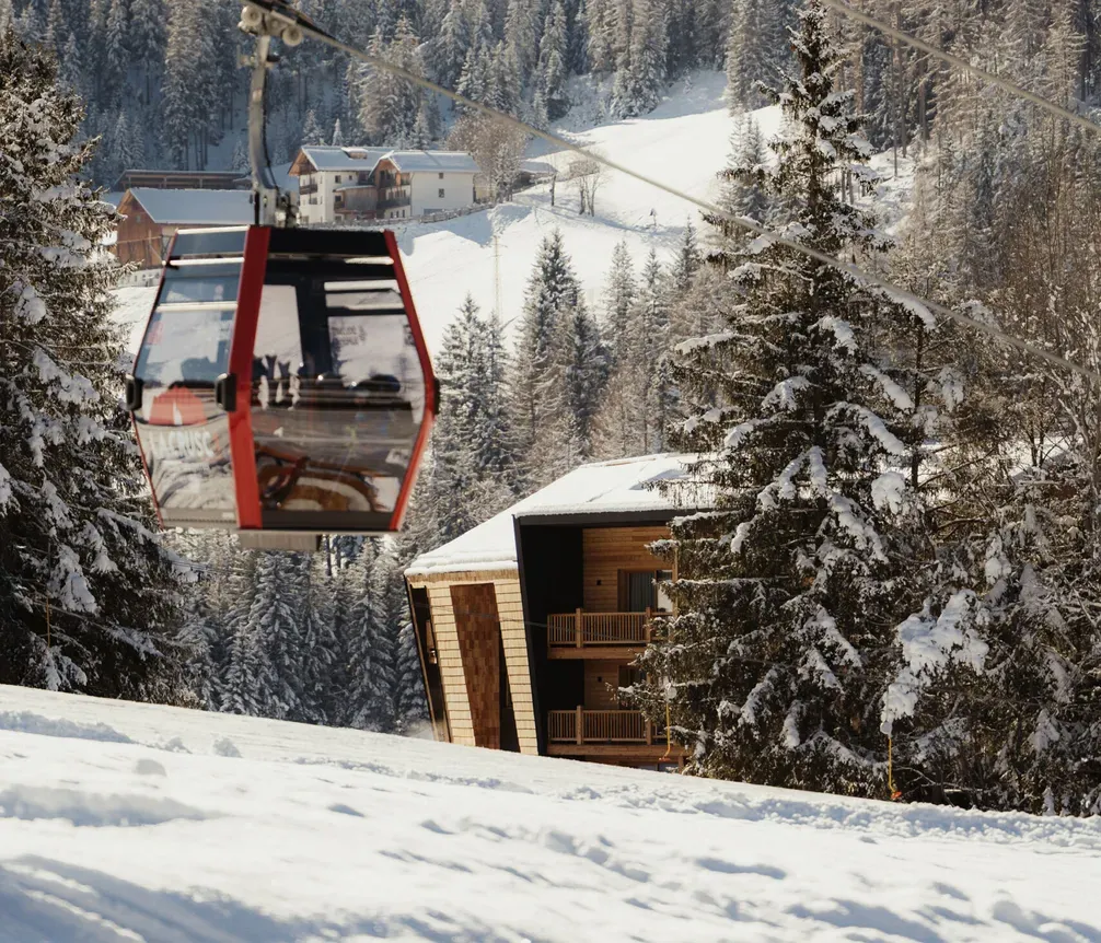 A red cable car glides over a snowy mountain landscape. Pine trees and cabins are nestled in the snow, with a clear blue sky above, evoking winter tranquility.