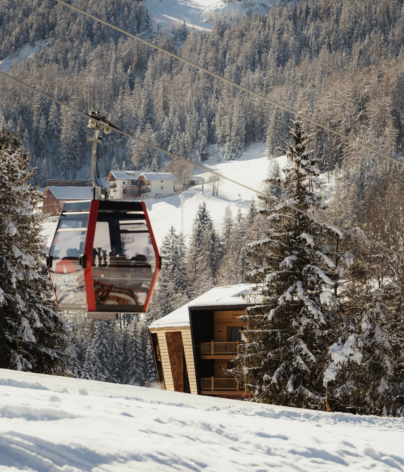 A red cable car glides over a snowy mountain landscape. Pine trees and cabins are nestled in the snow, with a clear blue sky above, evoking winter tranquility.