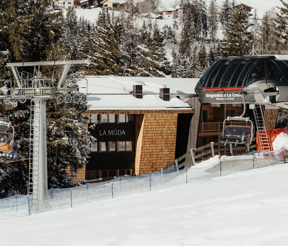 Ski resort scene with snowy slopes, pine trees, and modern gondolas. Log cabin labeled “La Müda” between chairlift stations against a wintery backdrop.