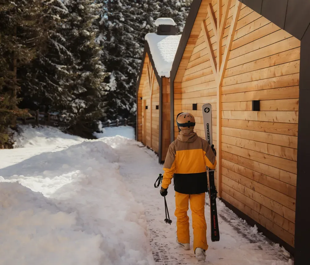 Person in yellow ski gear carrying skis walks alongside a wooden chalet in snowy woods. The scene is peaceful, with tall trees under a clear blue sky.