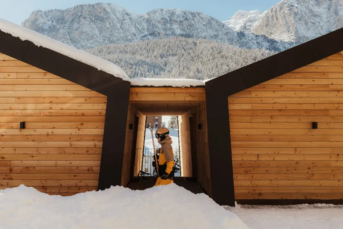A skier in yellow pants and a helmet walks through a snow-covered wooden structure, with majestic mountains in the background under a clear blue sky.