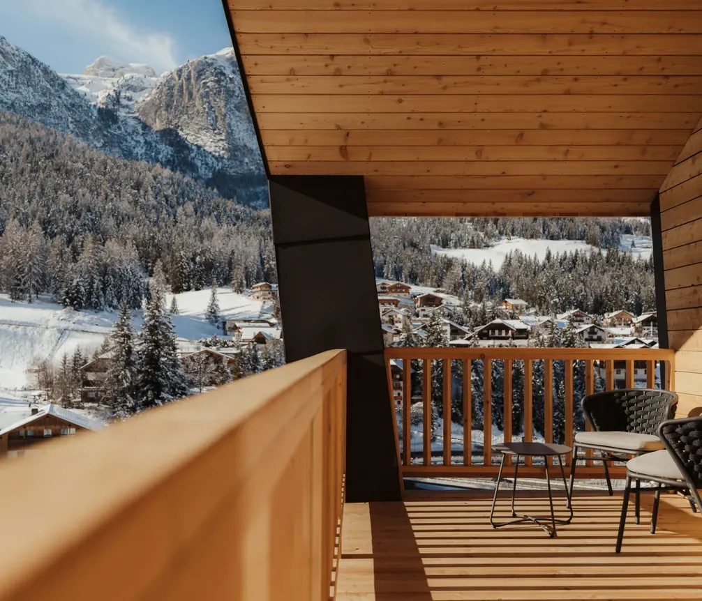 Sunny wooden balcony overlooking snowy mountains and pine trees. Two black chairs and a small table suggest cozy relaxation. Peaceful winter scene.