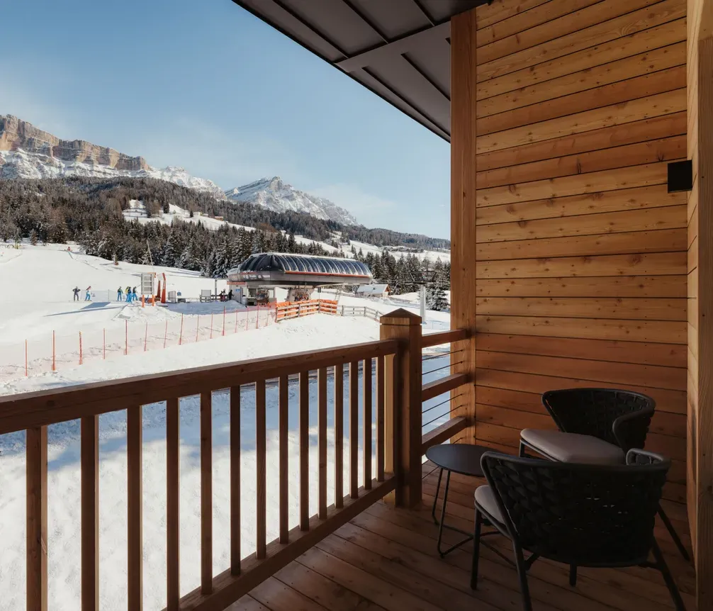 Cozy wooden balcony overlooking a snowy ski resort with mountain views. Skiers and a lift station are visible under a clear blue sky.