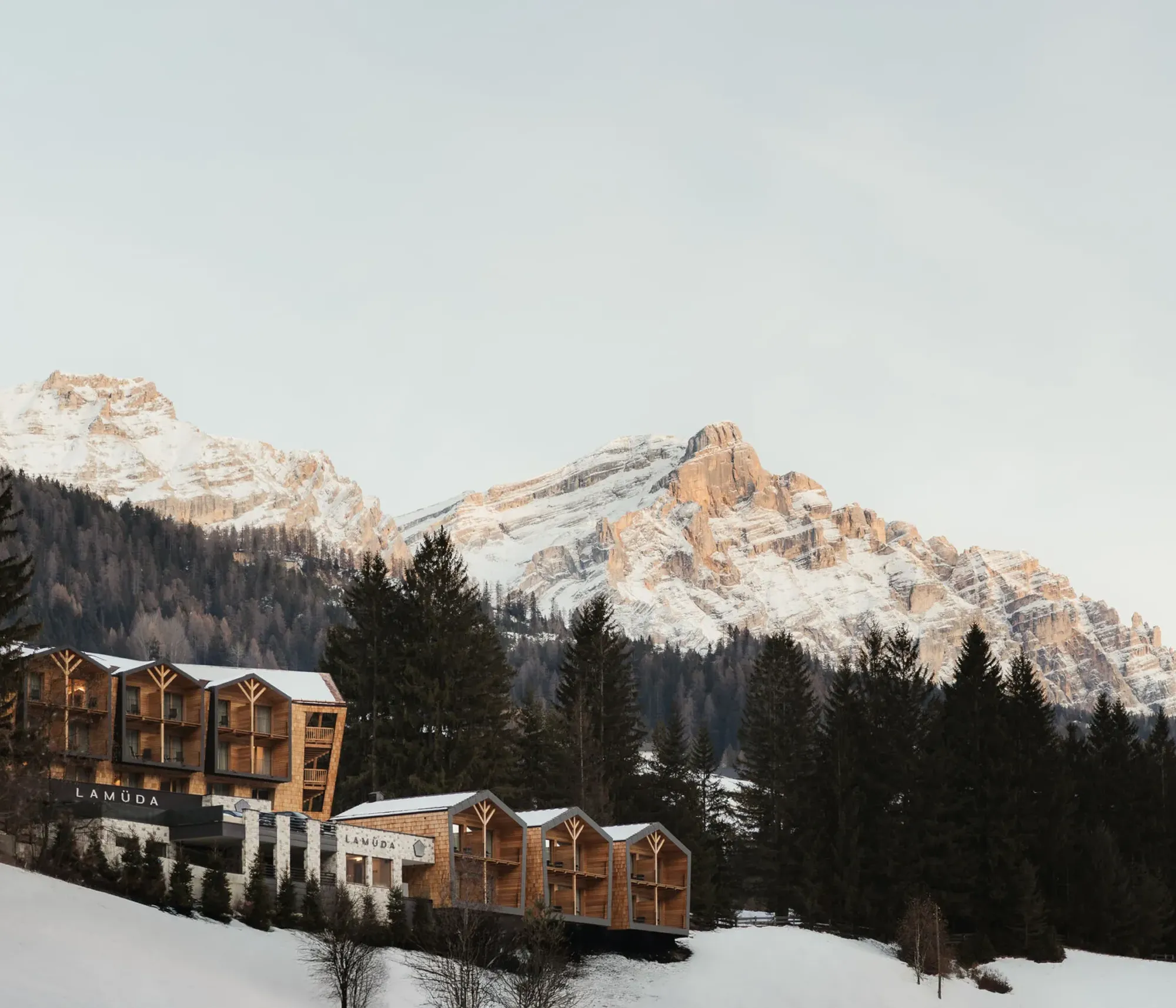Wooden cabins on a snowy hillside are surrounded by evergreen trees, with sunlit, rugged mountains in the background under a clear sky.