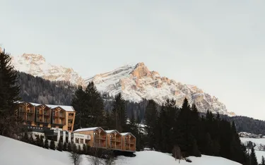 Wooden cabins on a snowy hillside are surrounded by evergreen trees, with sunlit, rugged mountains in the background under a clear sky.