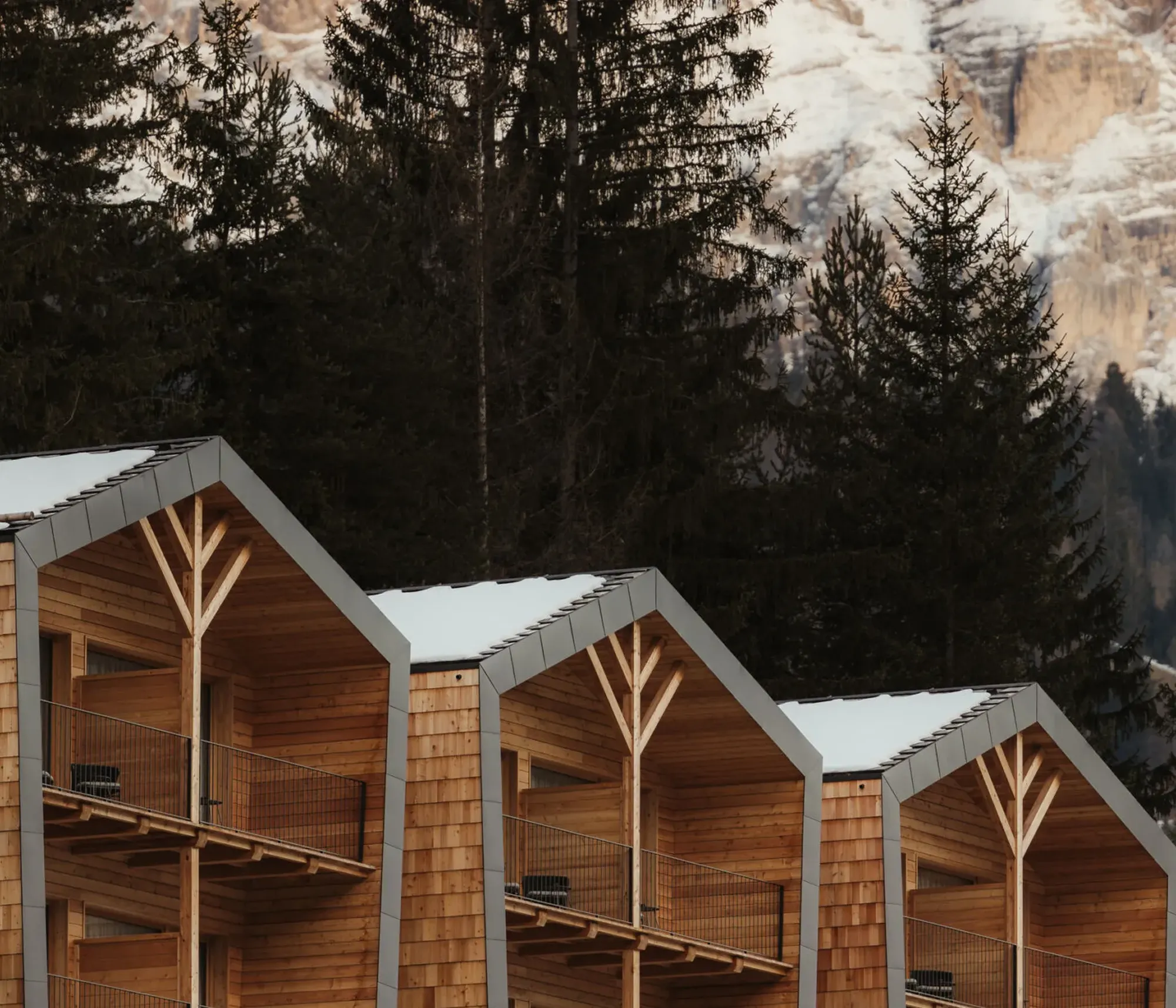 Modern wooden cabins with triangular roofs are nestled among tall, dark pine trees. Snow-capped mountains are illuminated by soft, warm sunlight.