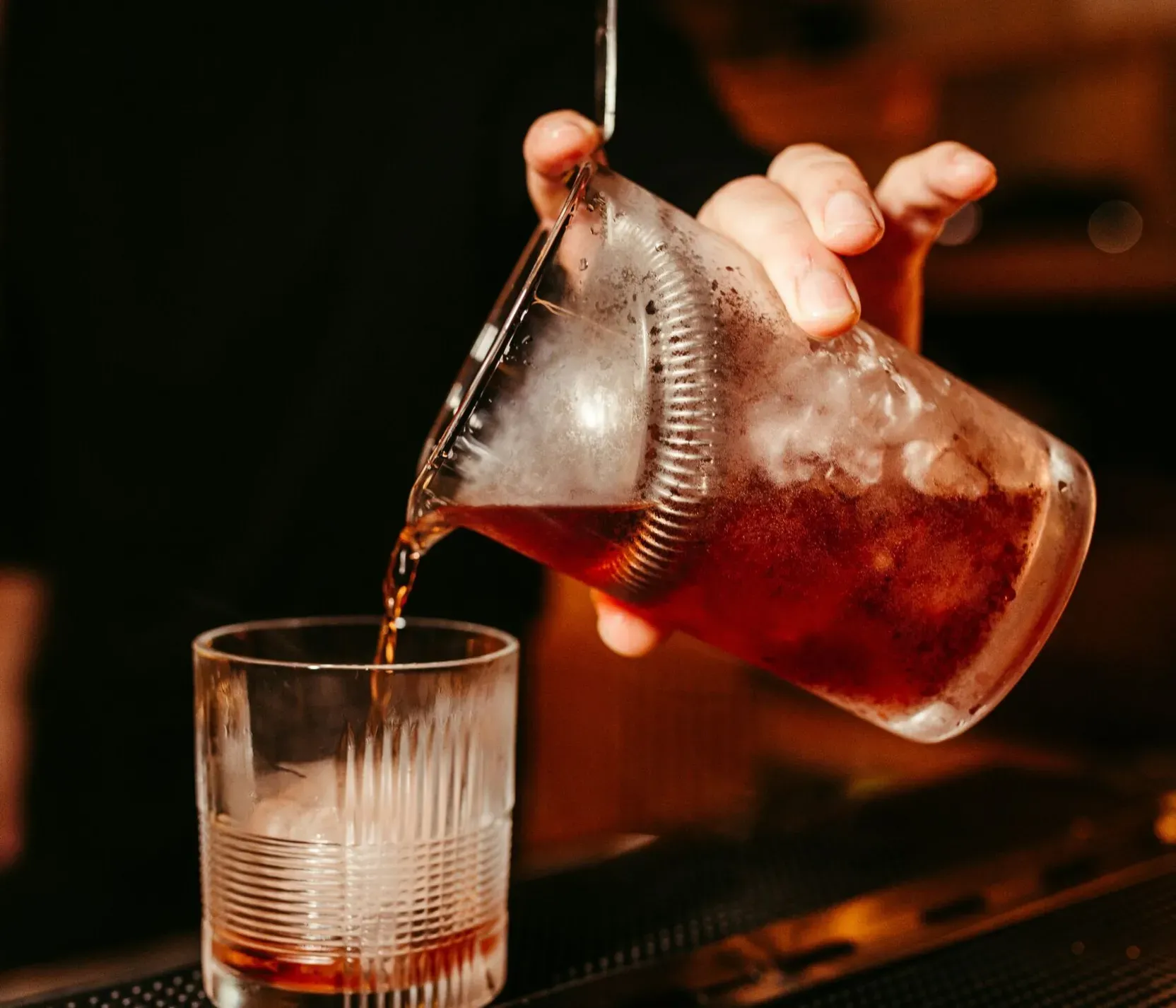 Ein Barkeeper gießt einen Getränk aus einem durchsichtigen Behälter in ein Glas. Der Hintergrund ist warm beleuchtet, Fokus liegt auf den Händen und dem Glas.