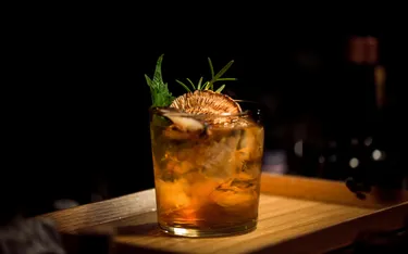 Ein Glas Whiskey mit Eis, garniert mit einer Ananas- und Rosmarin-Note, auf einem Holzbrett vor dunklem Hintergrund.
