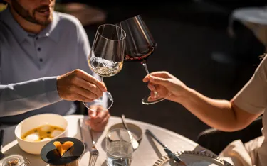 Zwei Personen stoßen mit Weingläsern an, während sie an einem festlich gedeckten Tisch mit Dessert, Wasser und Besteck sitzen.