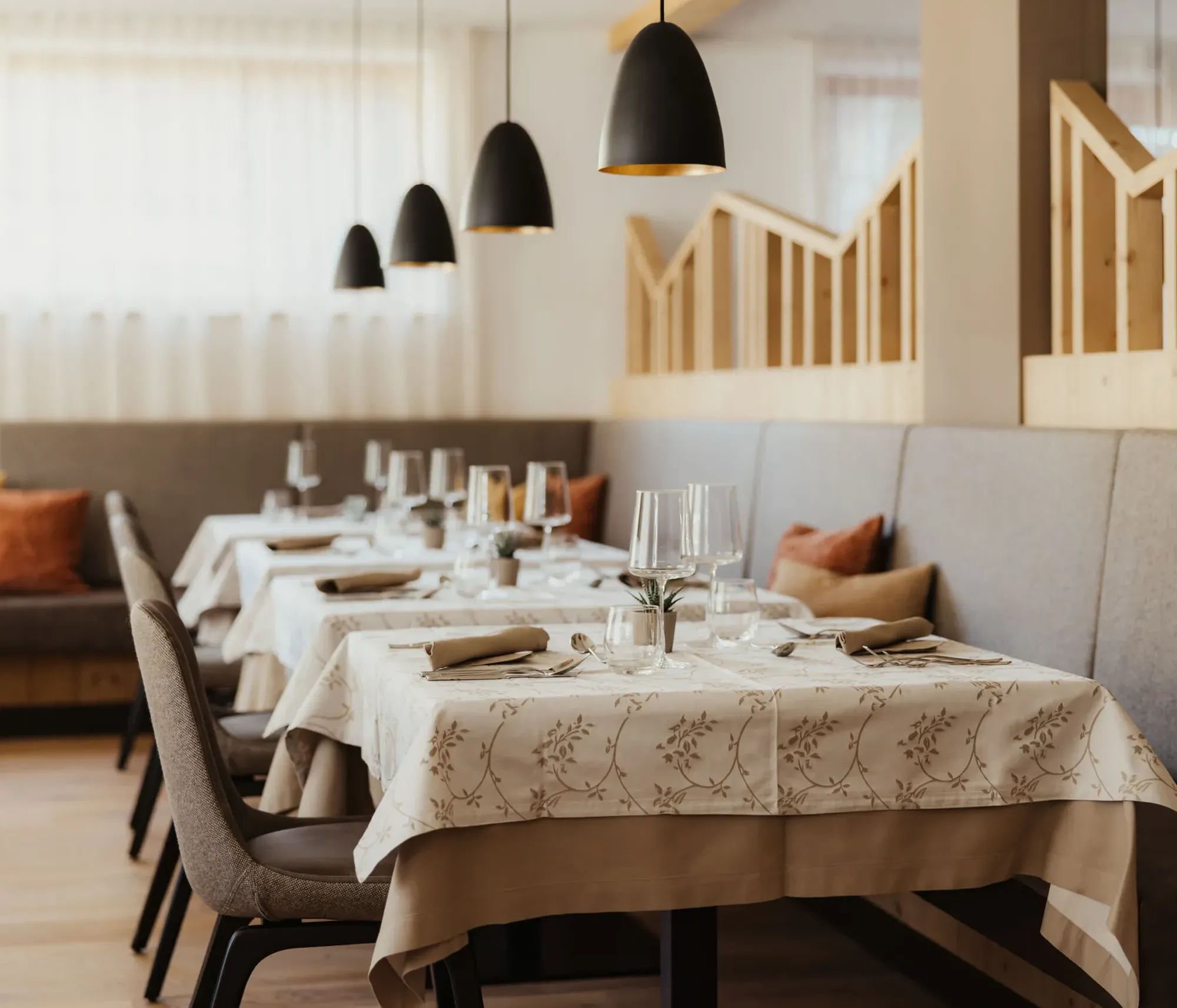Elegant restaurant interior with neatly set tables, white floral tablecloths, and soft lighting from black pendant lamps. Cozy, inviting atmosphere.