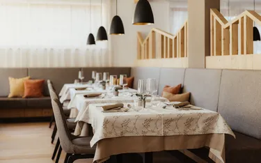 Elegantes Restaurantinterieur mit sorgfältig gedeckten Tischen, weißen Tischdecken mit Blumenmuster und sanfter Beleuchtung durch schwarze Pendelleuchten. Gemütliche, einladende Atmosphäre.