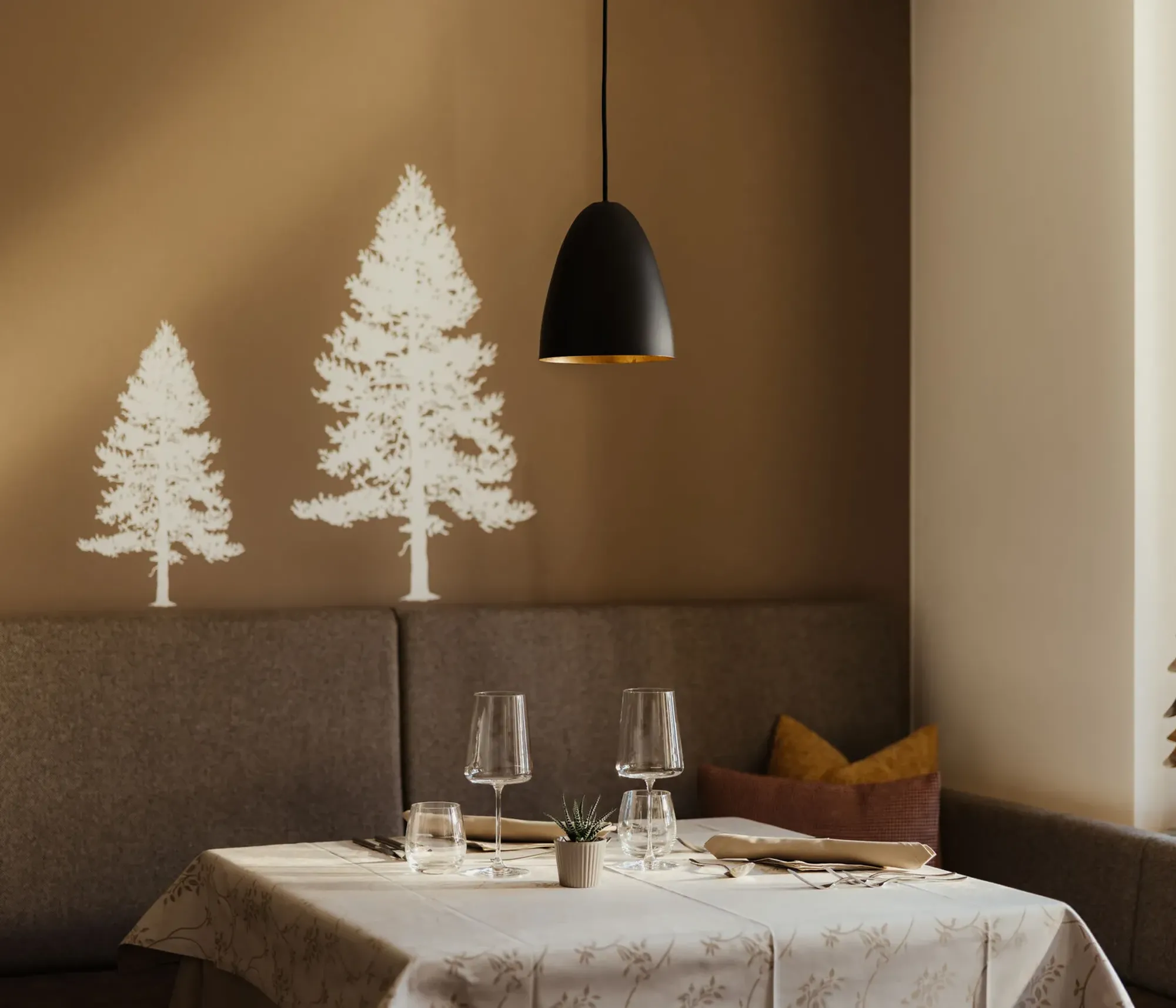 Cozy dining nook with brown walls featuring white tree decals. A table set for two with wine glasses and a plant centerpiece, lit by a black pendant lamp.