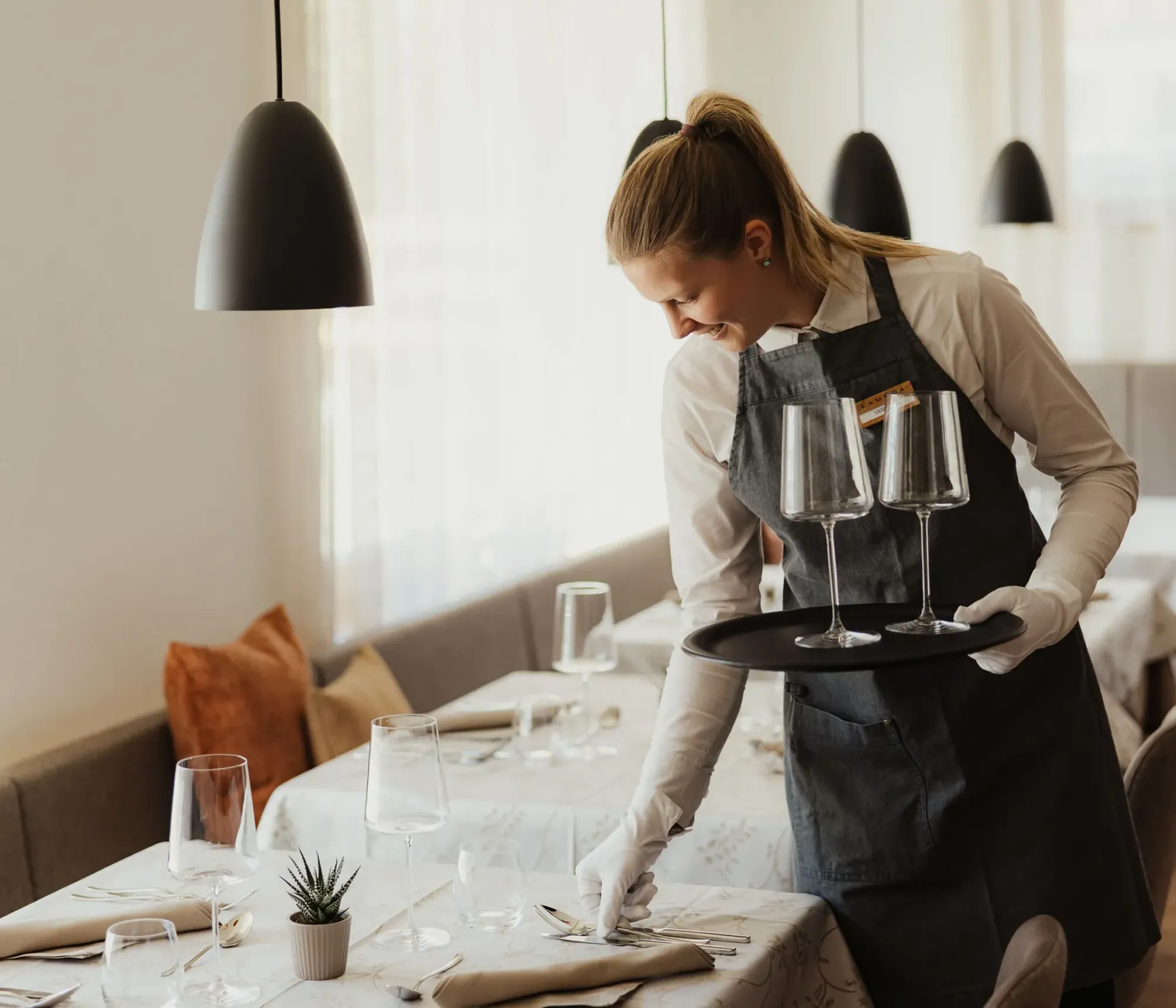 A smiling waitress in a gray apron sets a table with elegant glassware in a softly lit, cozy restaurant. The ambiance is warm and inviting.