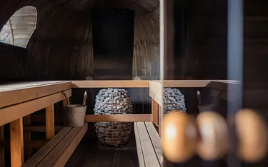 Innenraum einer Sauna mit Holzsitze und Steinen in einem Korb, dunkle Holzwände und ein kleines Fenster auf der linken Seite.