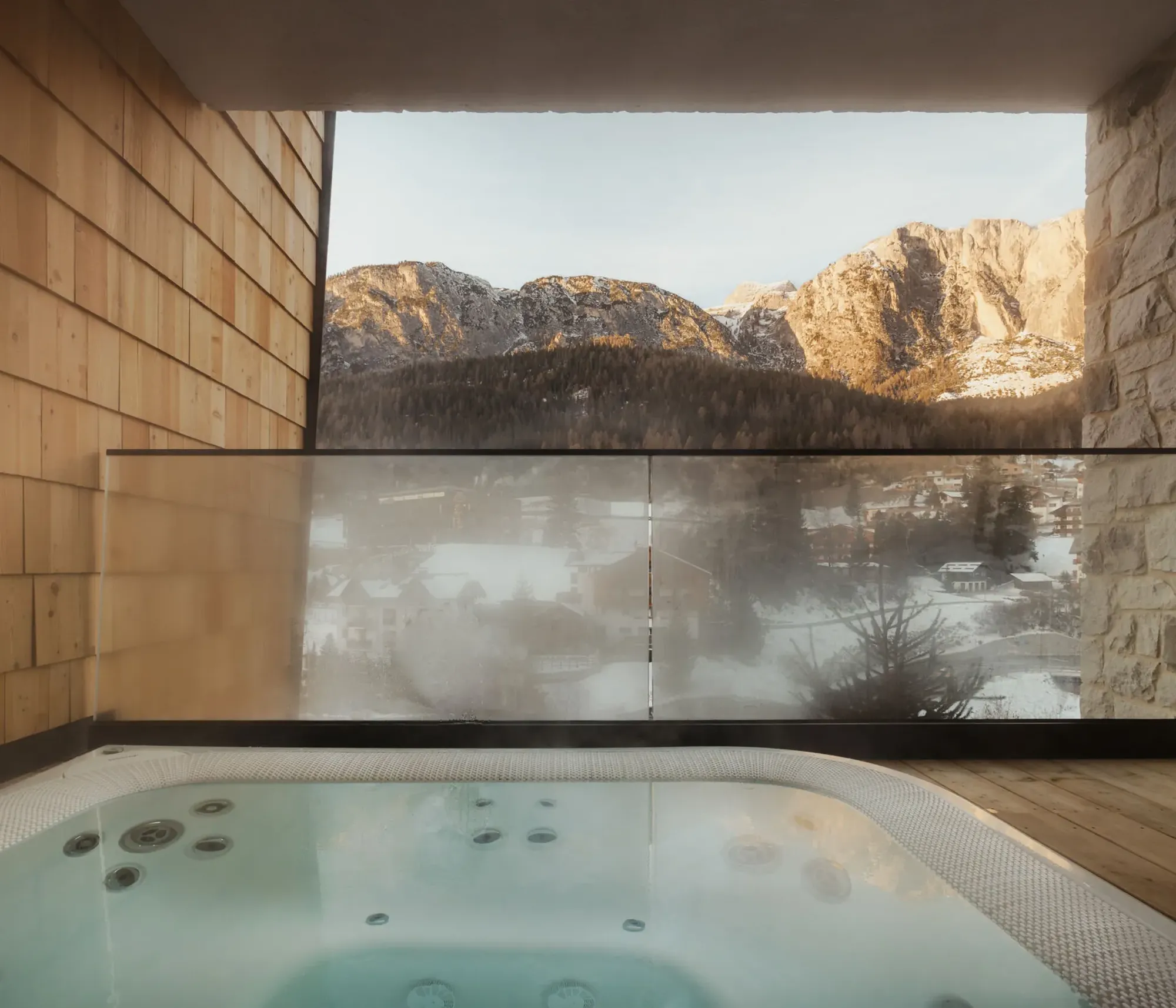 Rooftop hot tub with steaming water overlooks snow-covered mountain landscape. Wooden and stone walls add warmth and coziness to the serene view.