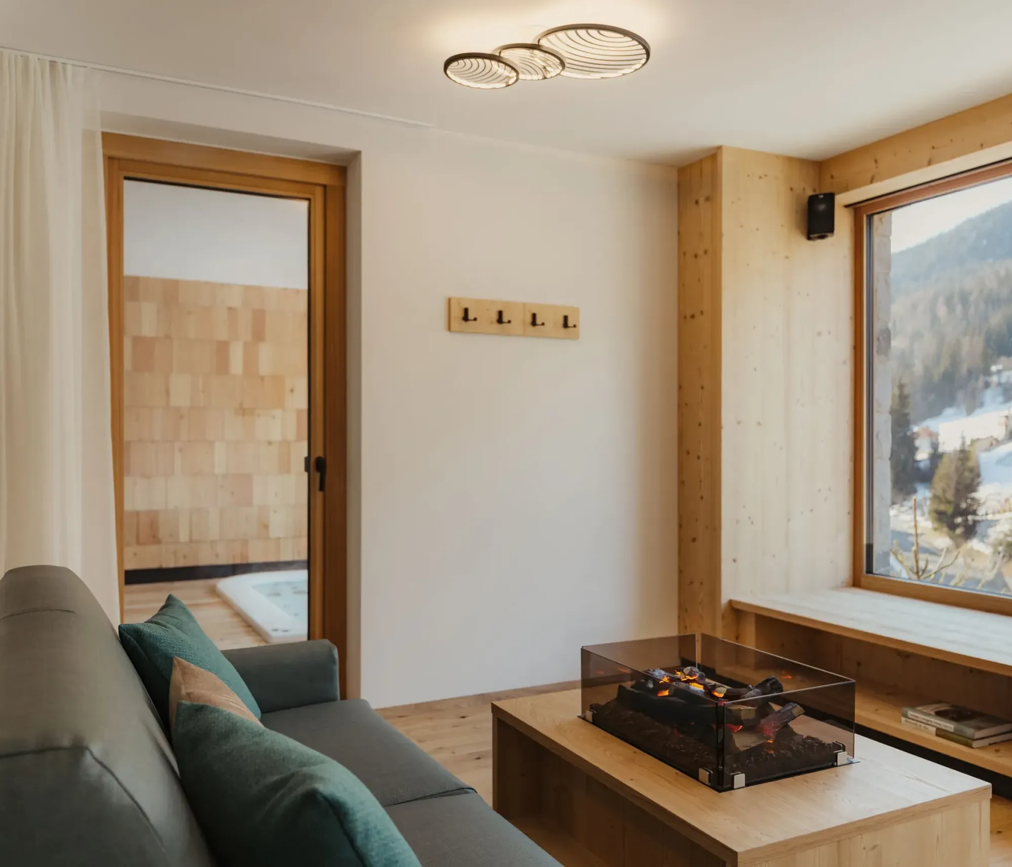 Cozy living room with large window overlooking snowy mountains. Modern decor with a green sofa, glass fireplace, and light wood accents. Peaceful ambiance.