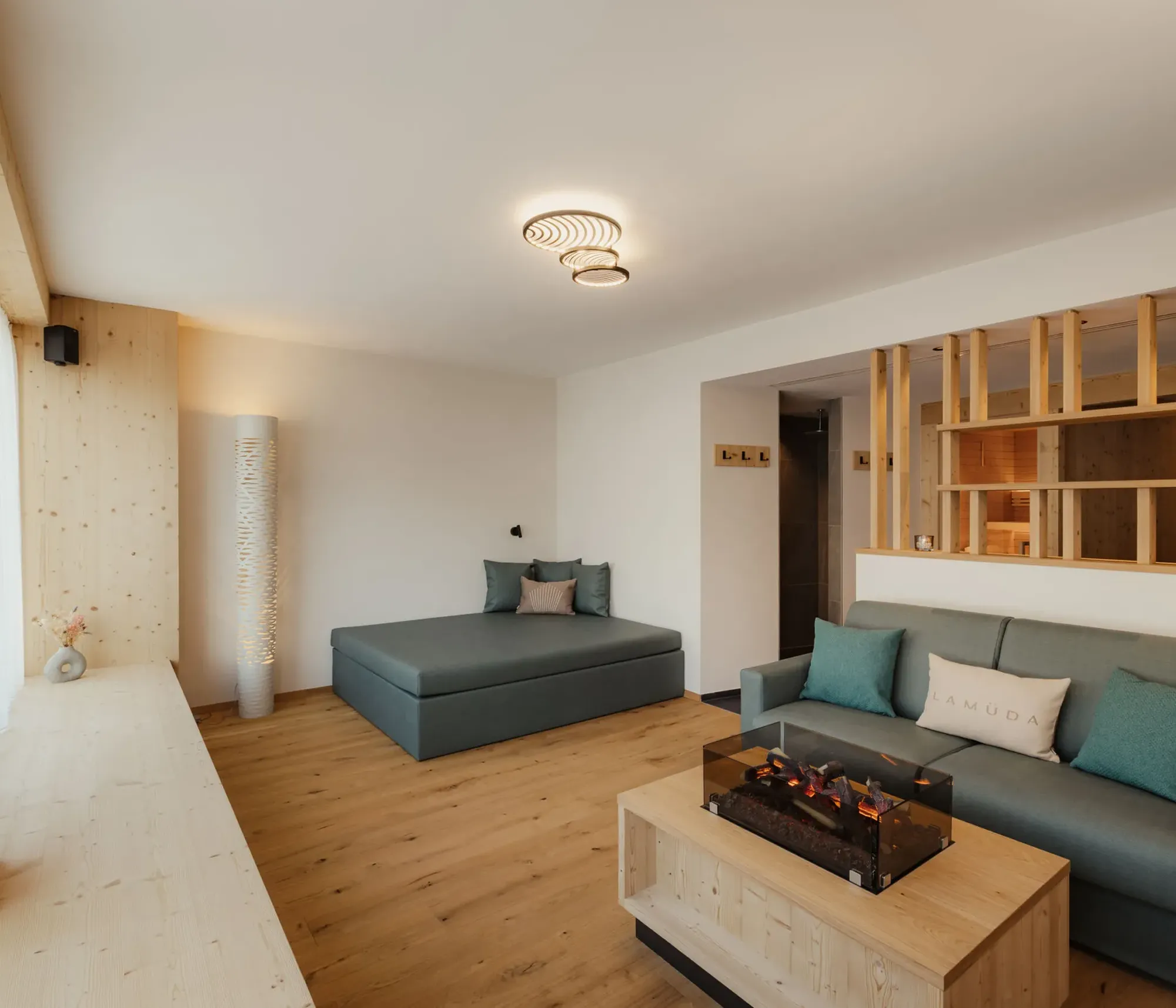 Modern living room with wooden floors and accents. Features a gray sofa, chaise lounge, and a coffee table with a faux fireplace. Soft, cozy ambiance.