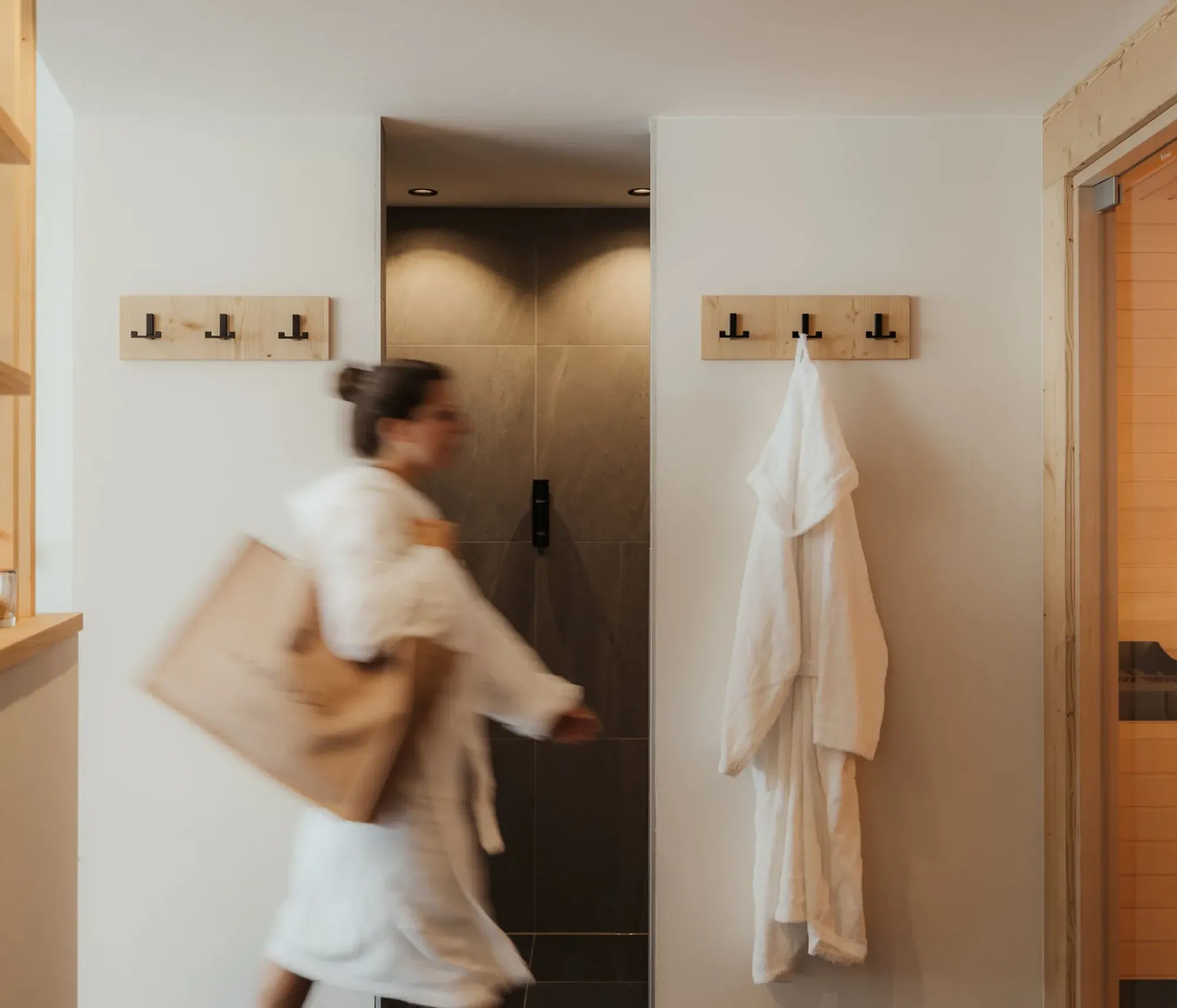 A person in a white robe walks through a modern spa hallway with wooden accents. A white robe hangs on a hook; a sauna room is visible on the right. Calm atmosphere.