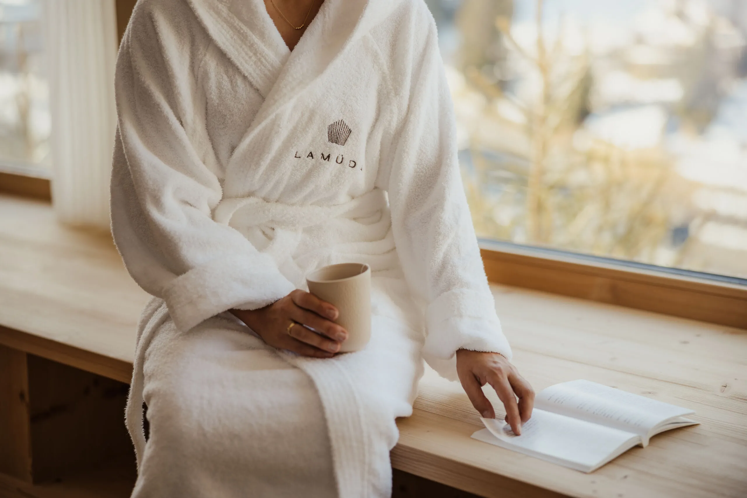 Eine Person in weißem Bademantel sitzt auf einer Holzbank am Fenster, hält eine Tasse und liest ein Buch. Die Szene vermittelt Ruhe und Wärme.