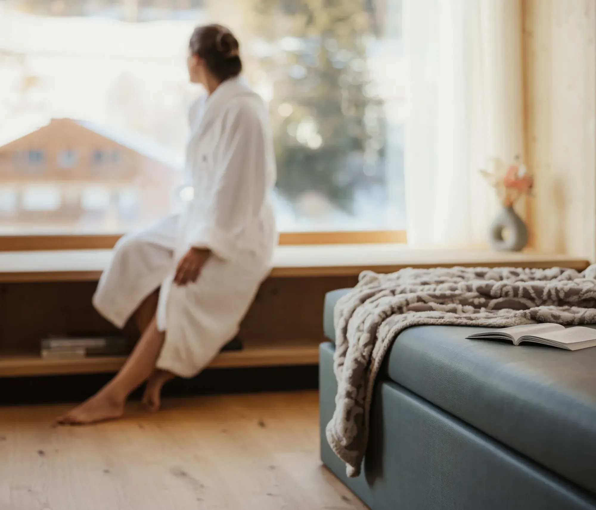 A person in a white robe sits by a large window, gazing outside at a snowy landscape. A cozy bed with a blanket and an open book is in the foreground.