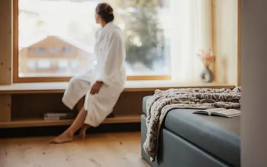 Eine Person in einem weißen Bademantel sitzt an einem großen Fenster und blickt auf die verschneite Landschaft draußen. Im Vordergrund befindet sich ein gemütliches Bett mit Decke und offenem Buch.