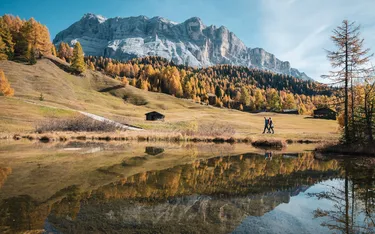 Berggipfel, bewaldete Hänge und eine klare Wasserfläche spiegeln die Landschaft in Alta Badia, Dolomiten, bei sonnigem Herbstwetter. Zwei Wanderer gehen am Ufer entlang.