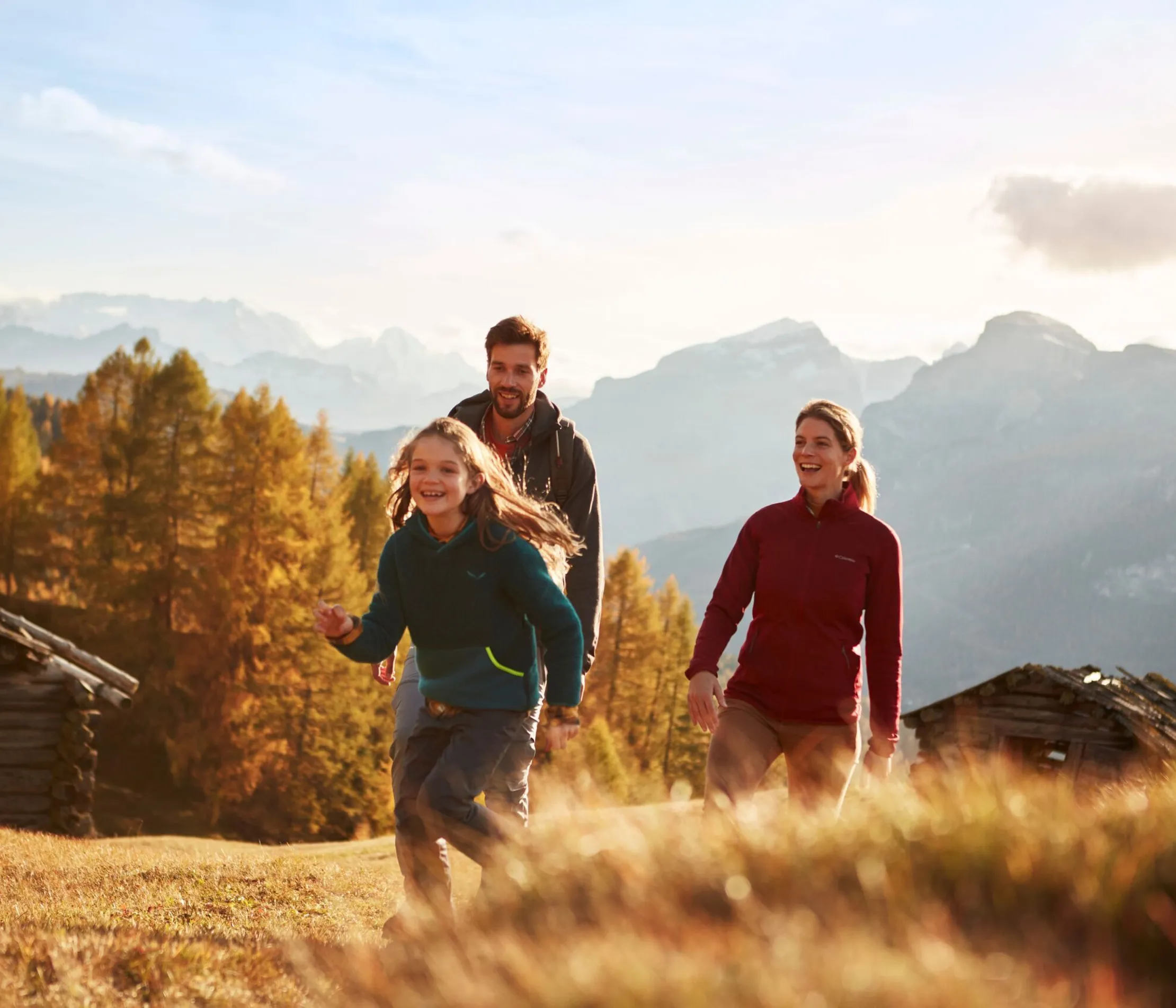 Famiglia che cammina in un paesaggio montano con alberi autunnali, montagne sullo sfondo e il cielo sereno sopra.