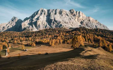 Berglandschaft in den Dolomiten mit orange- und grünfarbenen Bäumen, sanften Hügeln und kleinen Holzhütten unter einem blauen Himmel.