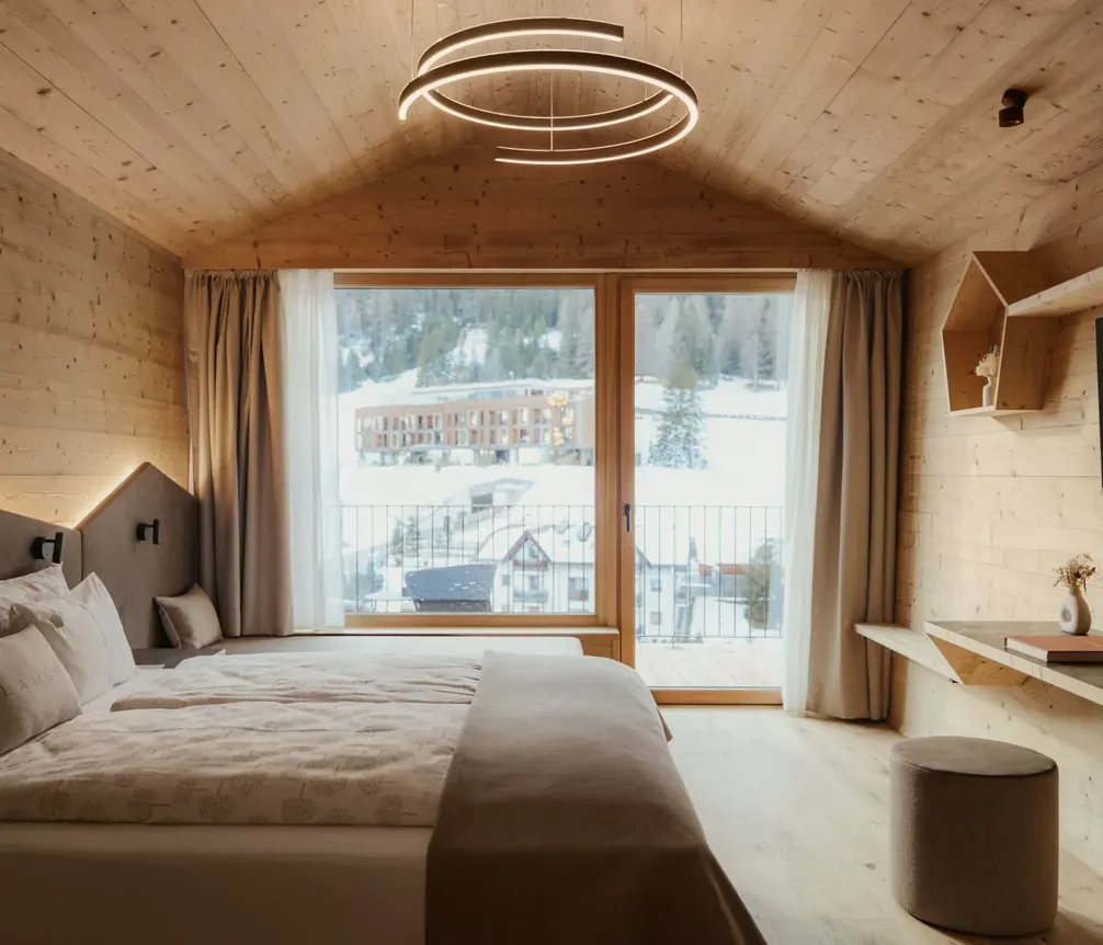 Cozy hotel room with wooden interiors and a large bed. A circular chandelier lights the space. Large window reveals a snowy mountain landscape outside.