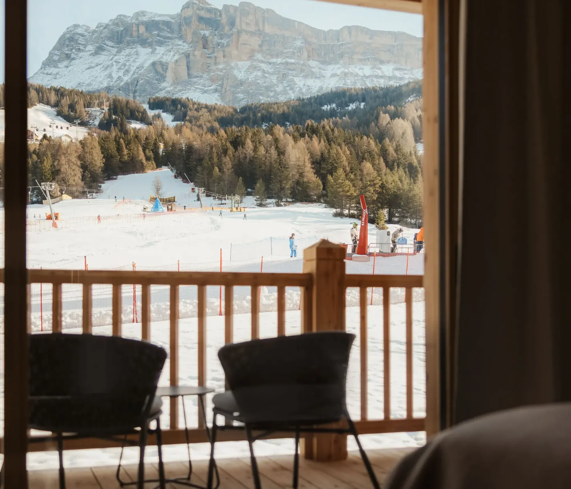 View from a cozy room showcasing a snowy ski resort scene with skiers, framed by a wooden balcony railing and distant snowy mountains.