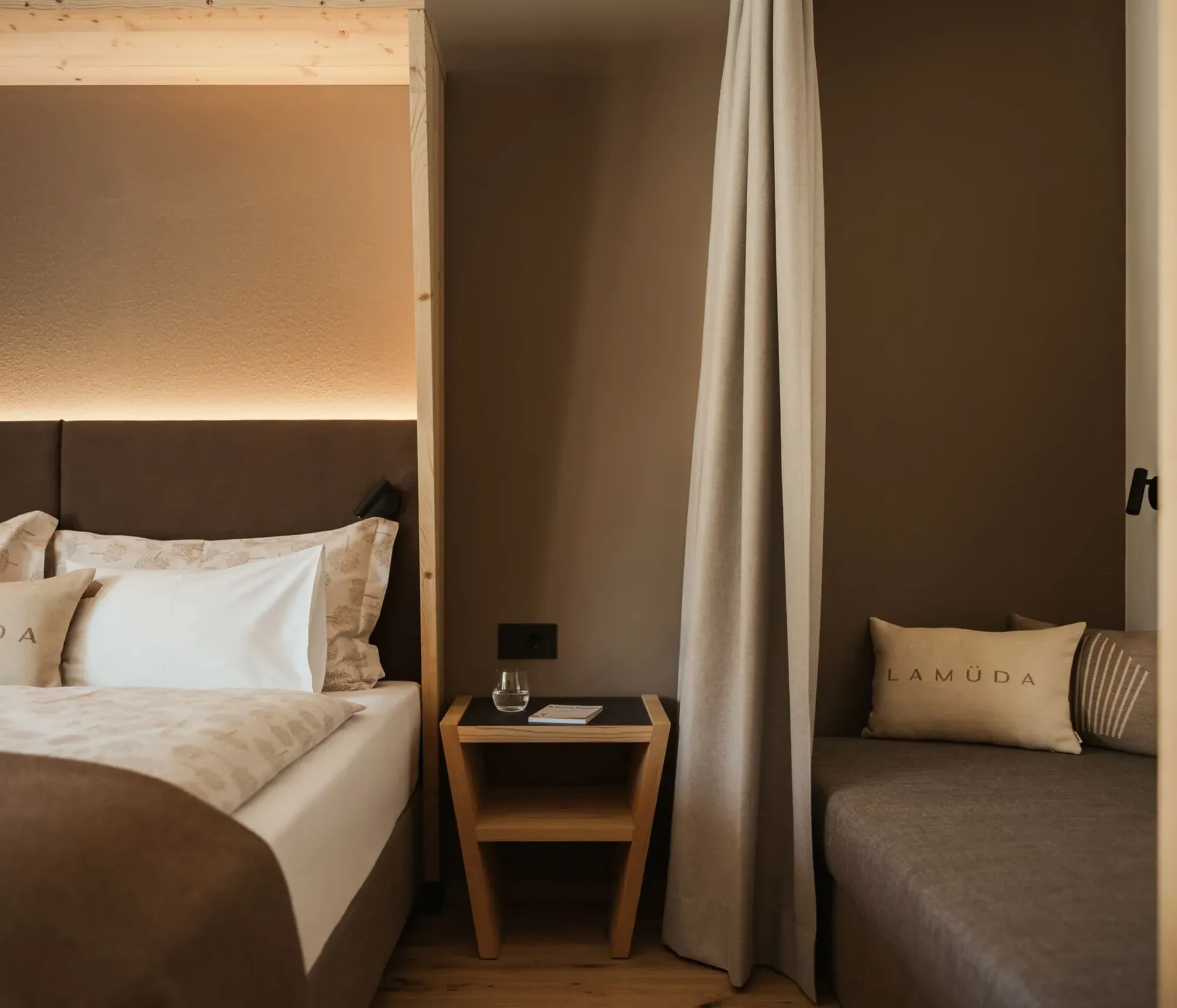 Cozy bedroom with a modern design, featuring a brown bed with labeled pillows, soft lighting, a wooden nightstand with a glass of water, and taupe curtains.