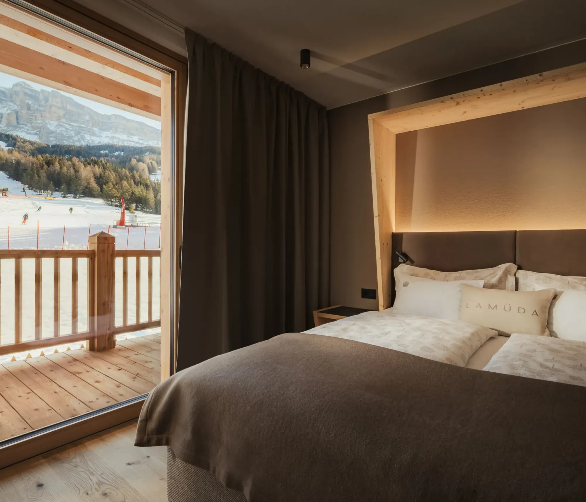 Cozy bedroom with a large bed, warm lighting, and a wooden frame. Balcony door reveals a snowy ski slope with forested mountains, creating a tranquil atmosphere.