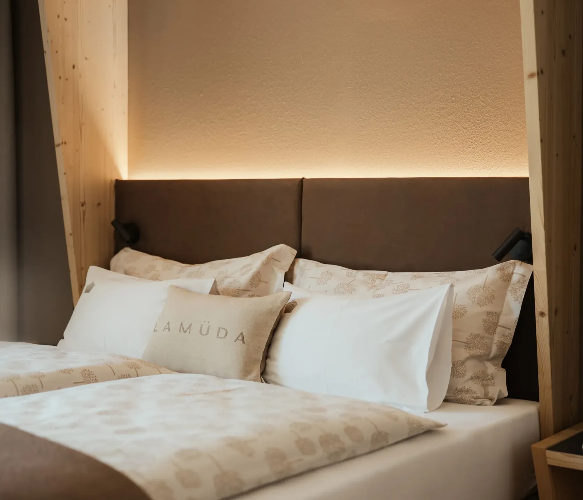 Cozy bedroom with a soft, brown headboard, illuminated by warm lighting. Neatly arranged pillows and a beige duvet create a calming ambiance.