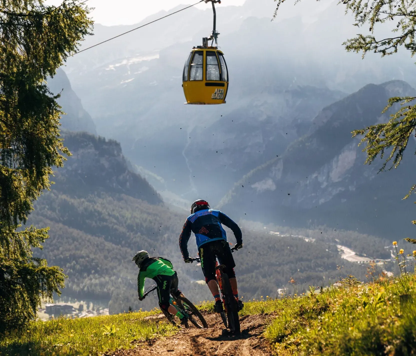 Two mountain bikers riding down a dirt trail in a lush green landscape with mountains in the background, while a yellow cable car moves above them in Alta Badia.