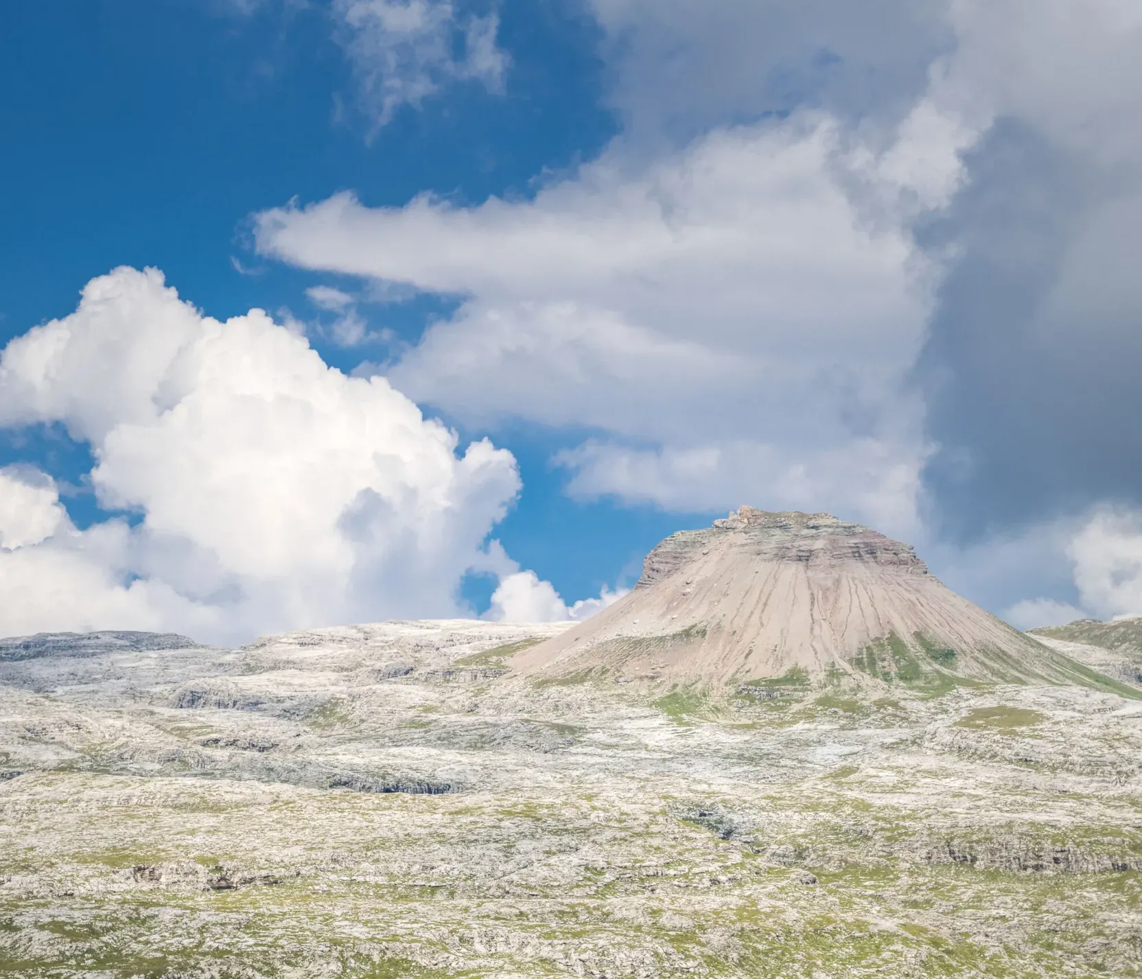 Paesaggio montano con nuvole bianche e cielo blu, rotto da una cima rocciosa e landa di rocce e erba verdi.