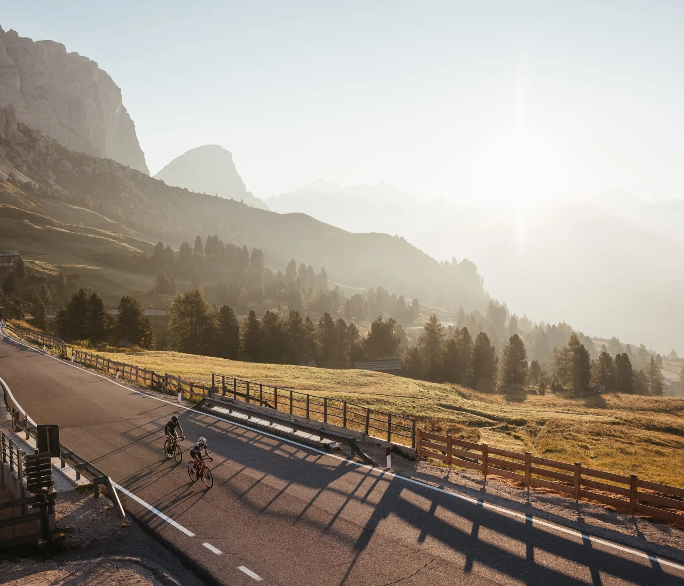 Scenic mountain landscape with a winding road, cyclists riding, lush green fields, and forested hills under bright sunlight in Alta Badia, Dolomites.